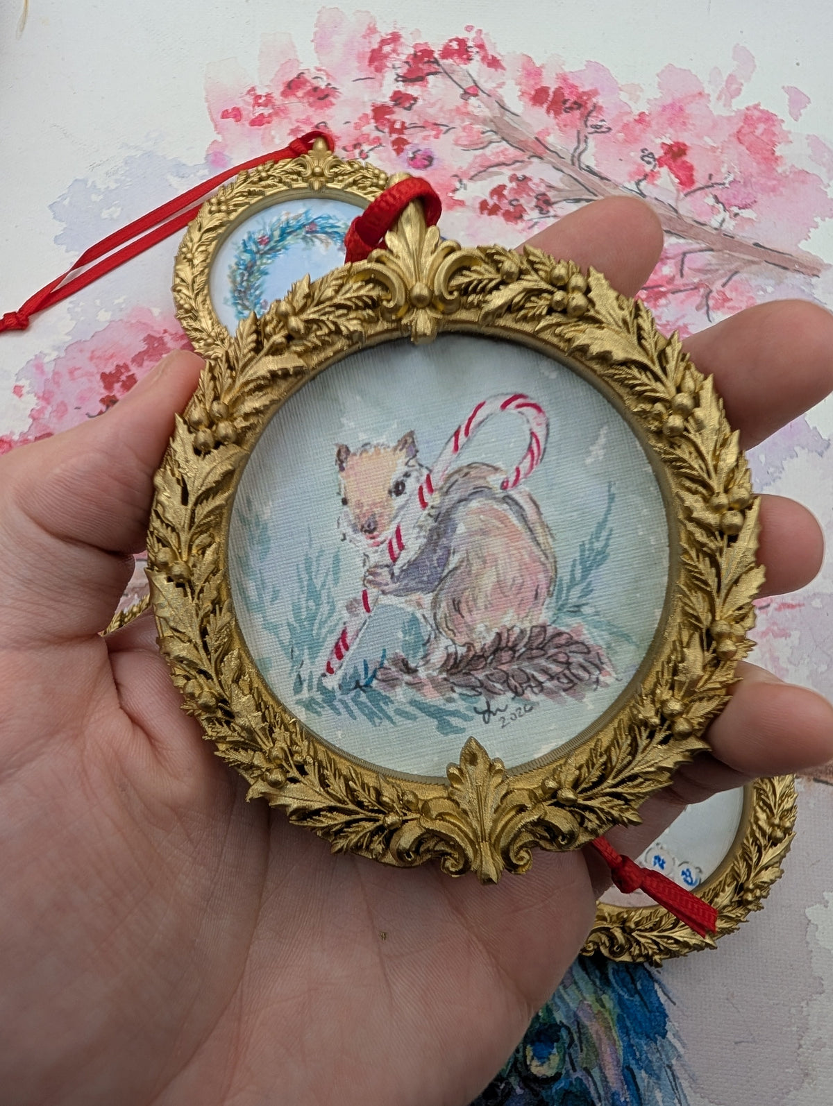 Chipmunk with a Candy Cane watercolor ornament in a gold wreath frame, held in hand